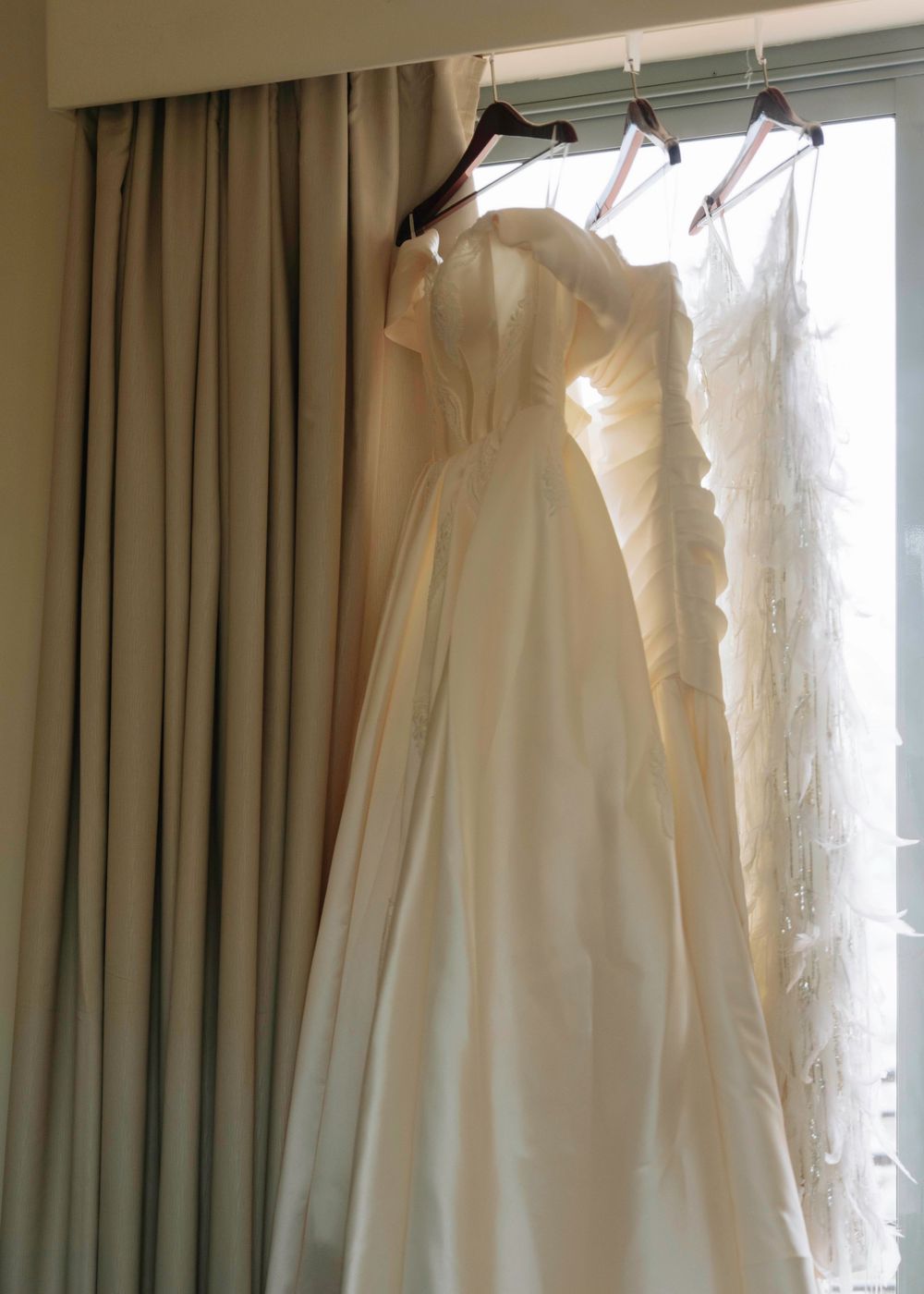 White wedding gowns hanging by sunlit window with flowing silhouettes.