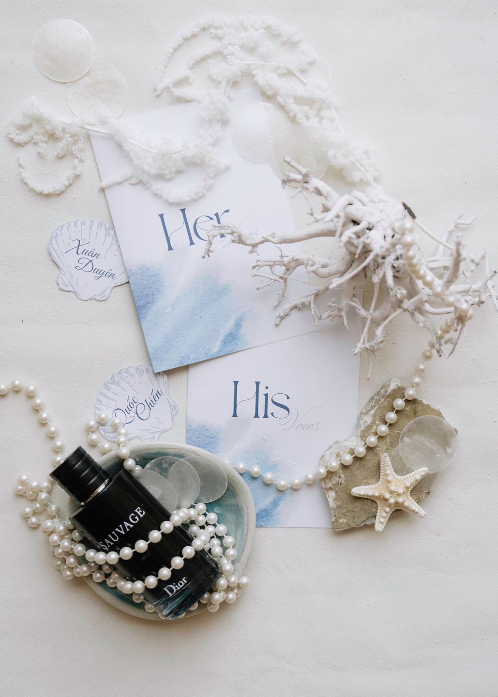 Flat lay of wedding vows cards, pearl necklace, Dior perfume, and coastal decor.