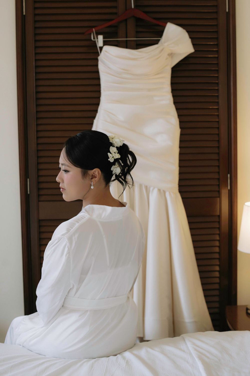 Bride in white robe sitting on bed gazing at her elegant mermaid wedding gown hanging nearby.