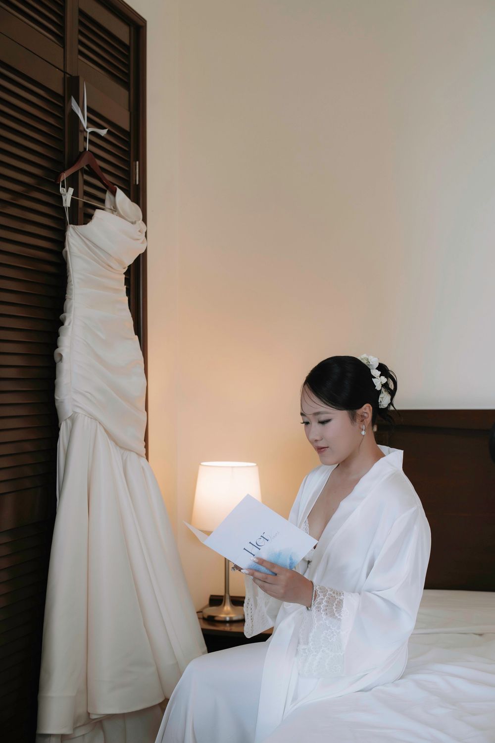 Bride in white robe reading a letter beside her hanging wedding gown in a warmly lit room.