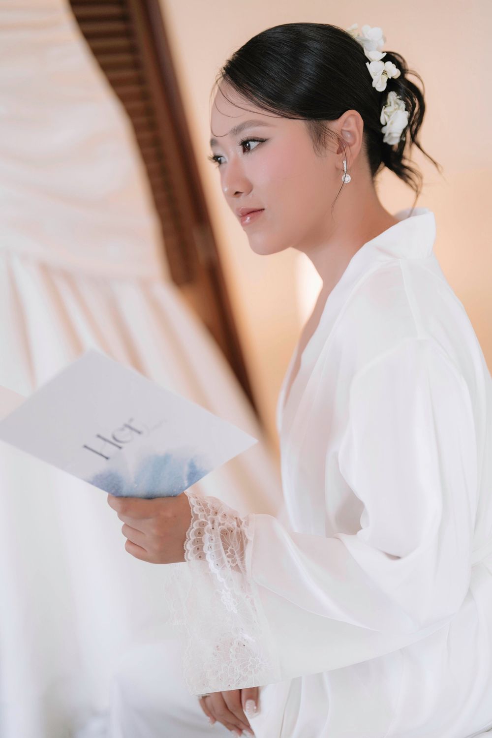Bride in white lace robe reading wedding vows card with white flower hair accessory.