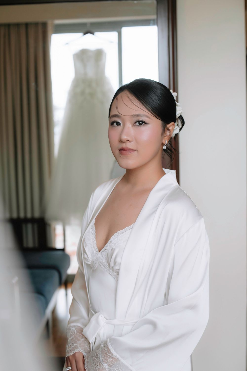 Beautiful Asian bride in white satin robe with wedding dress hanging in background.