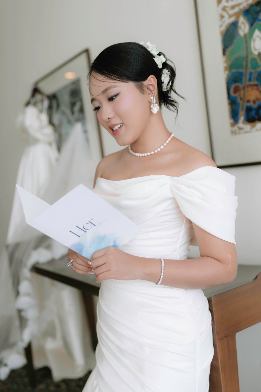 Smiling Asian bride in off-shoulder white gown reading heartfelt wedding vows letter.