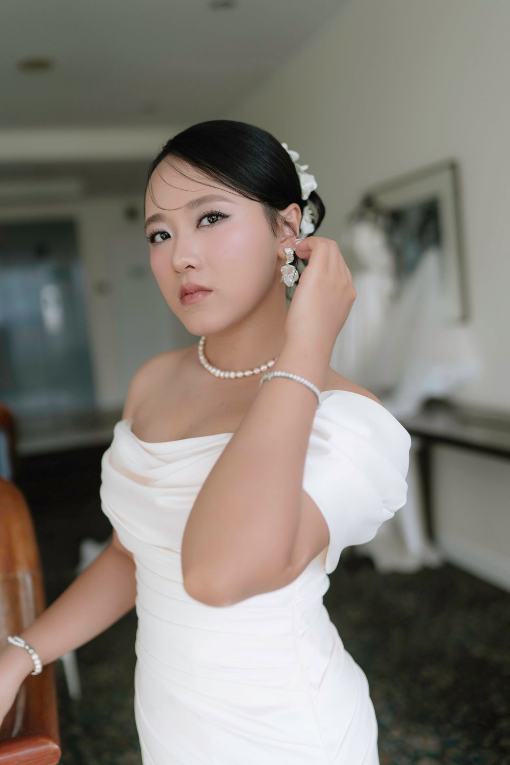 Asian bride in white mermaid gown adjusting pearl earring with elegant updo hairstyle.