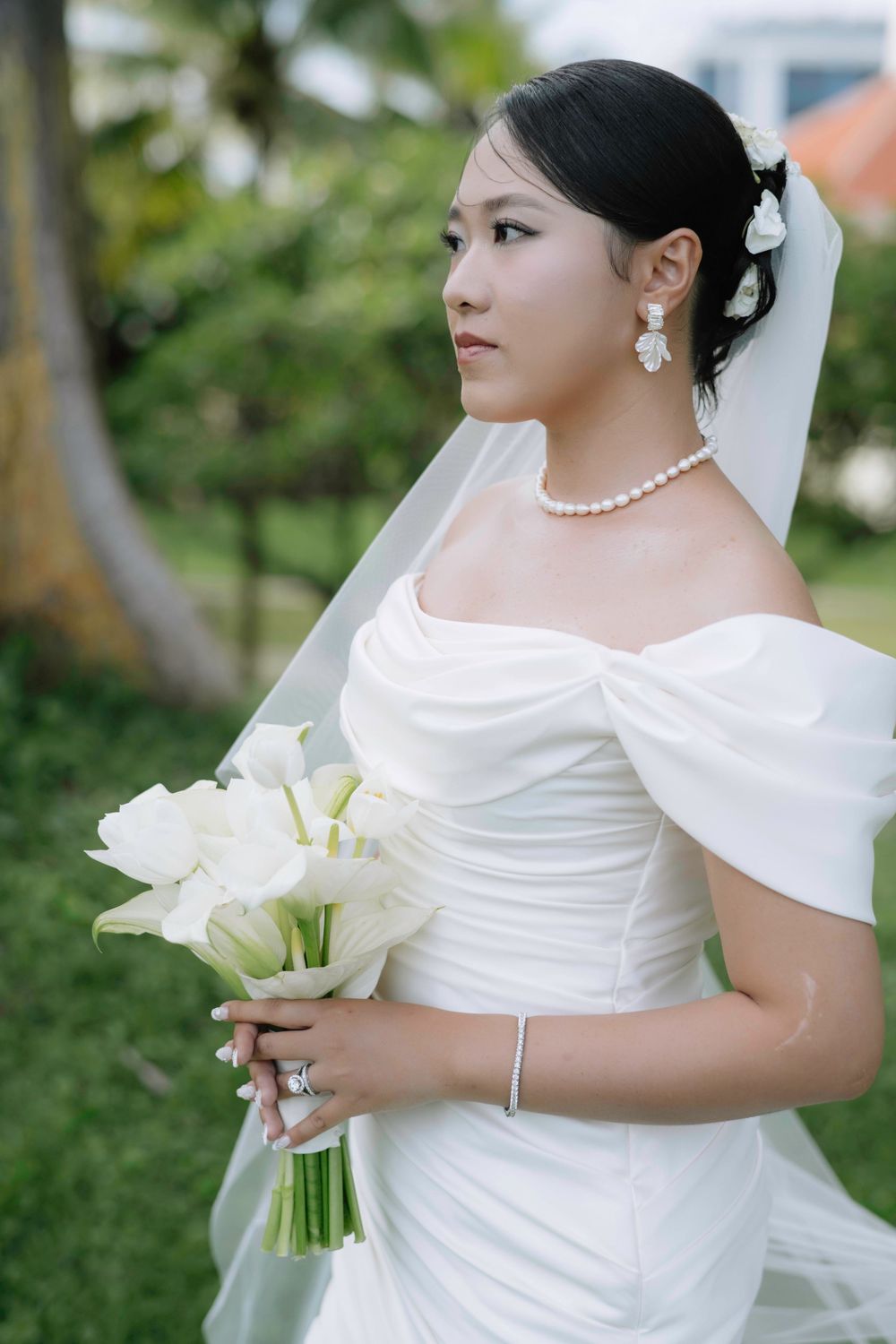 Asian bride in white off-shoulder wedding gown holding calla lily bouquet with pearl jewelry.