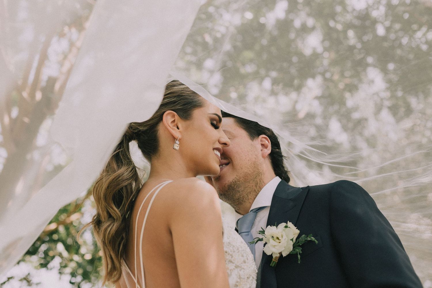 Romantic wedding moment captured under a flowing white veil in an outdoor setting.