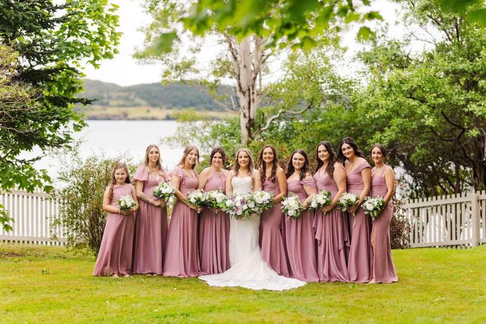Wedding party in mauve dresses and gray suits posed in front of white picket fence and green landscape.
