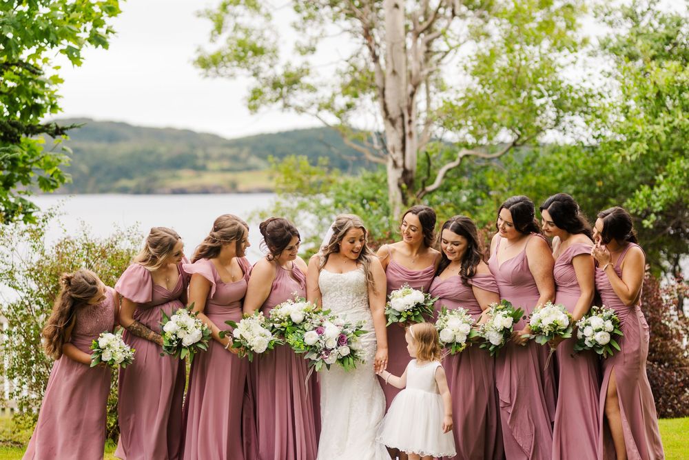 A group of bridesmaids in dusty pink and mauve dresses create a coordinated lineup at an outdoor lakeside wedding ceremony.
