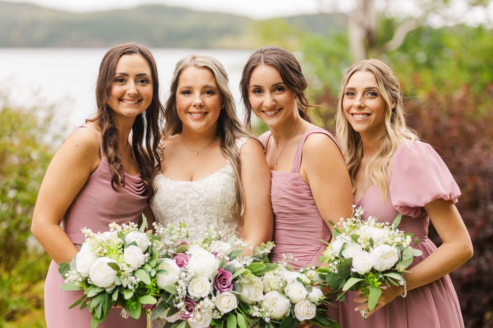 A group wearing dusty rose bridesmaid dresses hold white floral bouquets while posing together outdoors.