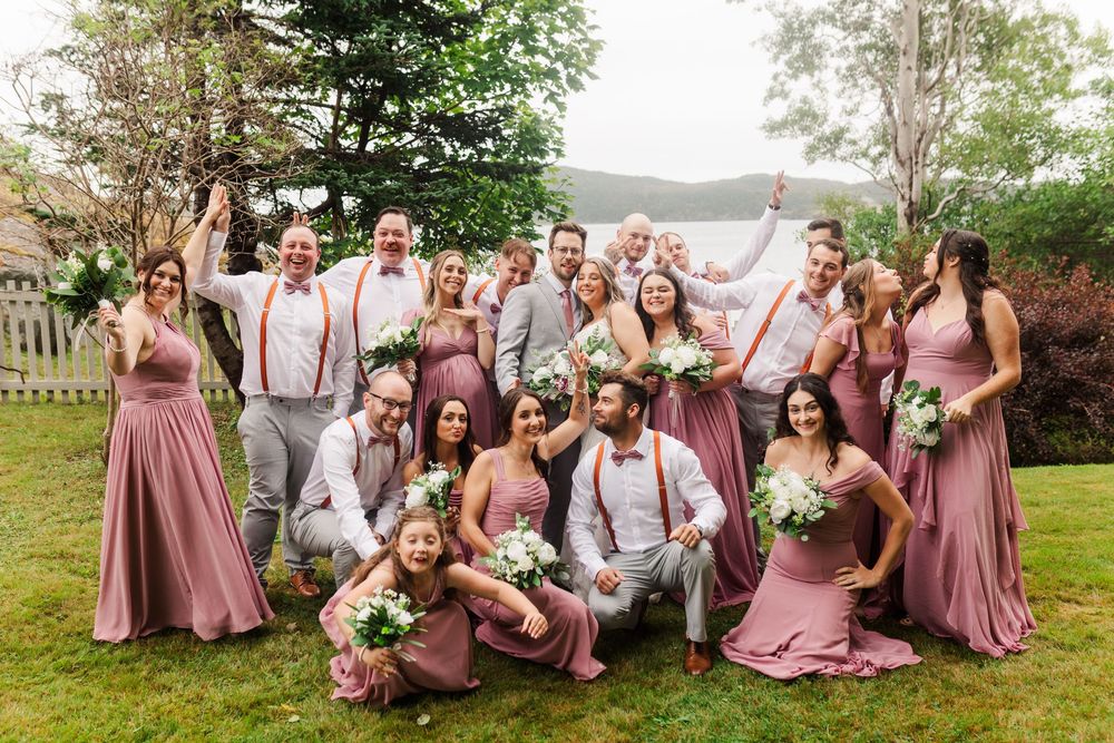 Wedding party in mauve dresses and red suspenders poses playfully together on grassy hillside for outdoor photos.