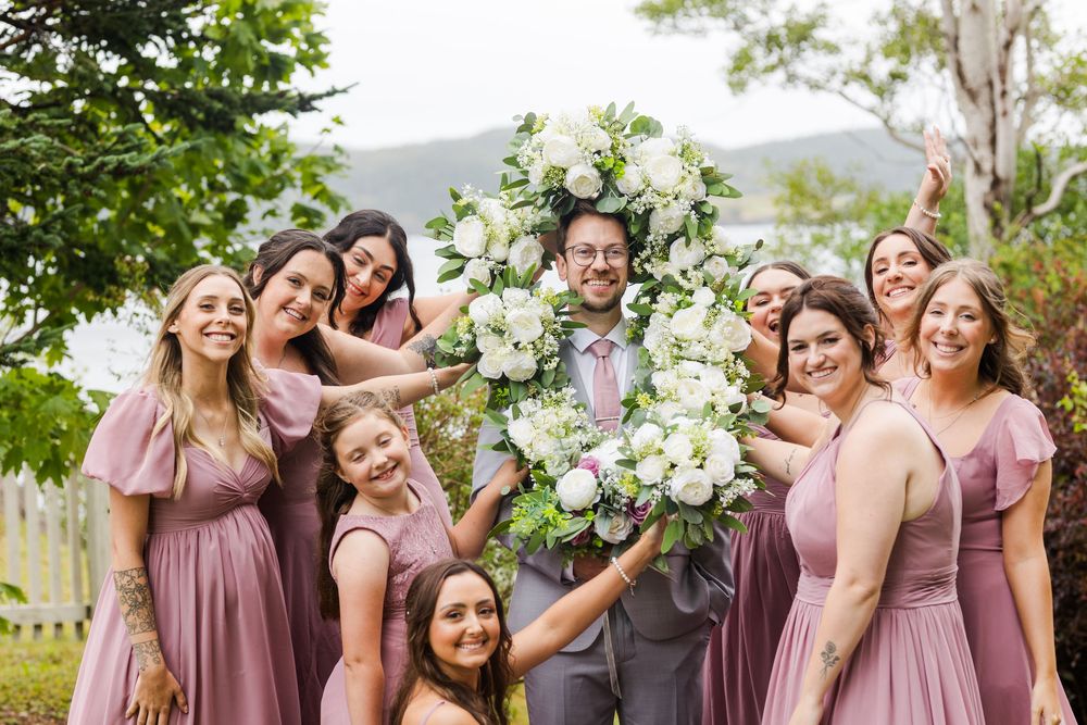 Wedding party in dusty rose dresses laughs and poses together while holding white floral bouquets outdoors.