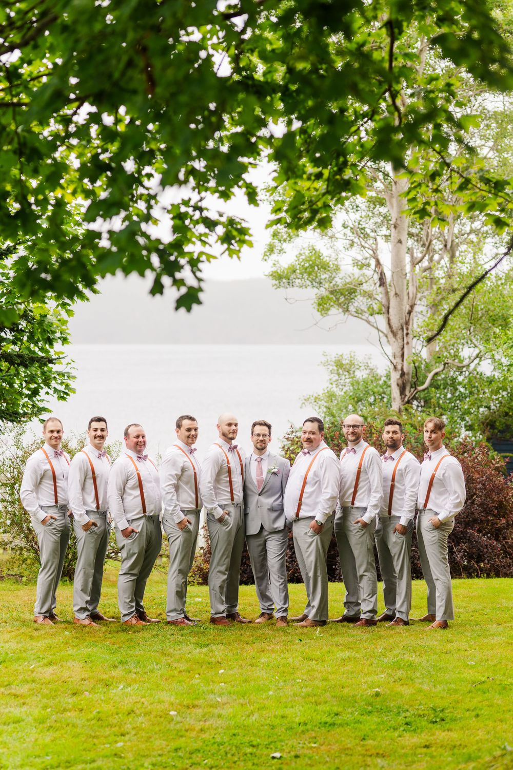 A line of groomsmen in matching gray suits and suspenders pose on a grassy lawn under green trees.