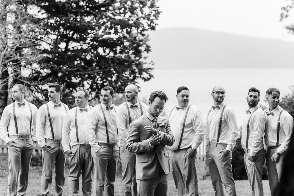 Groomsmen in light gray suits and suspenders stand together under a shady tree for formal photos.