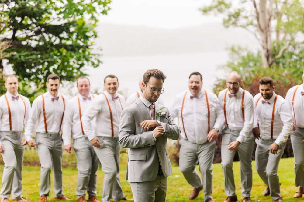 Wedding party wearing gray suits and pink suspenders pose playfully together outdoors in spring garden setting.