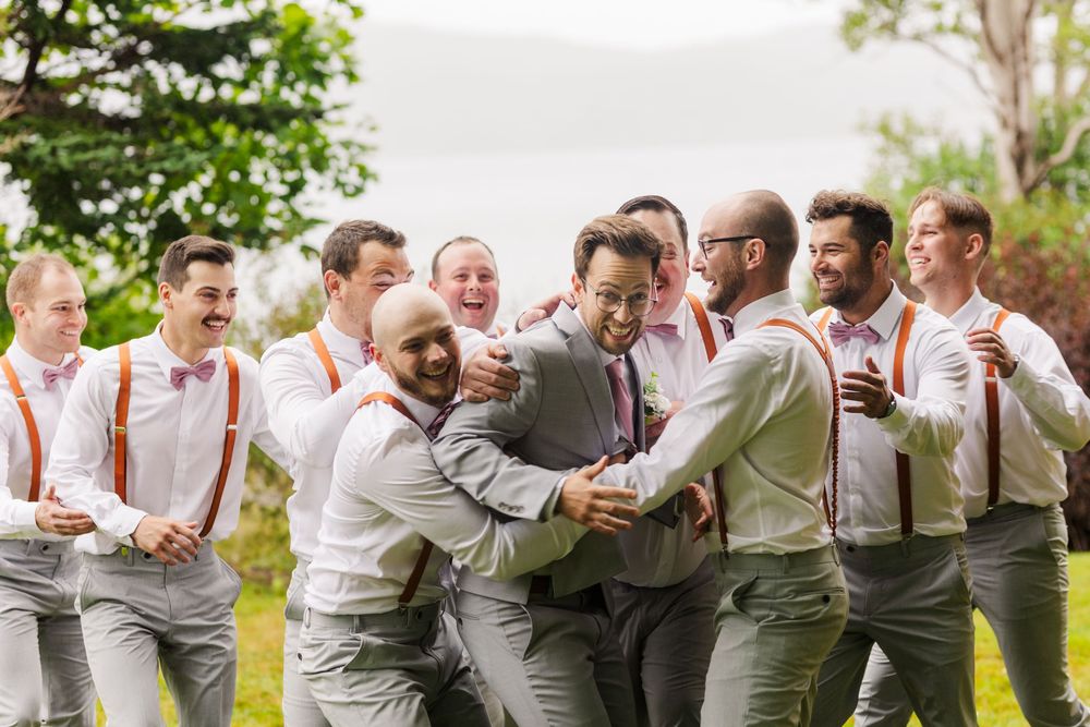 Groomsmen lined up in casual poses wearing gray pants and white shirts for a rustic outdoor wedding.