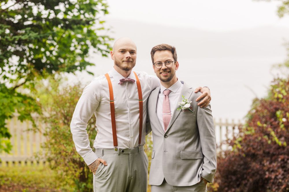 A group in gray suits and coral suspenders gather casually on a lawn for wedding photos.