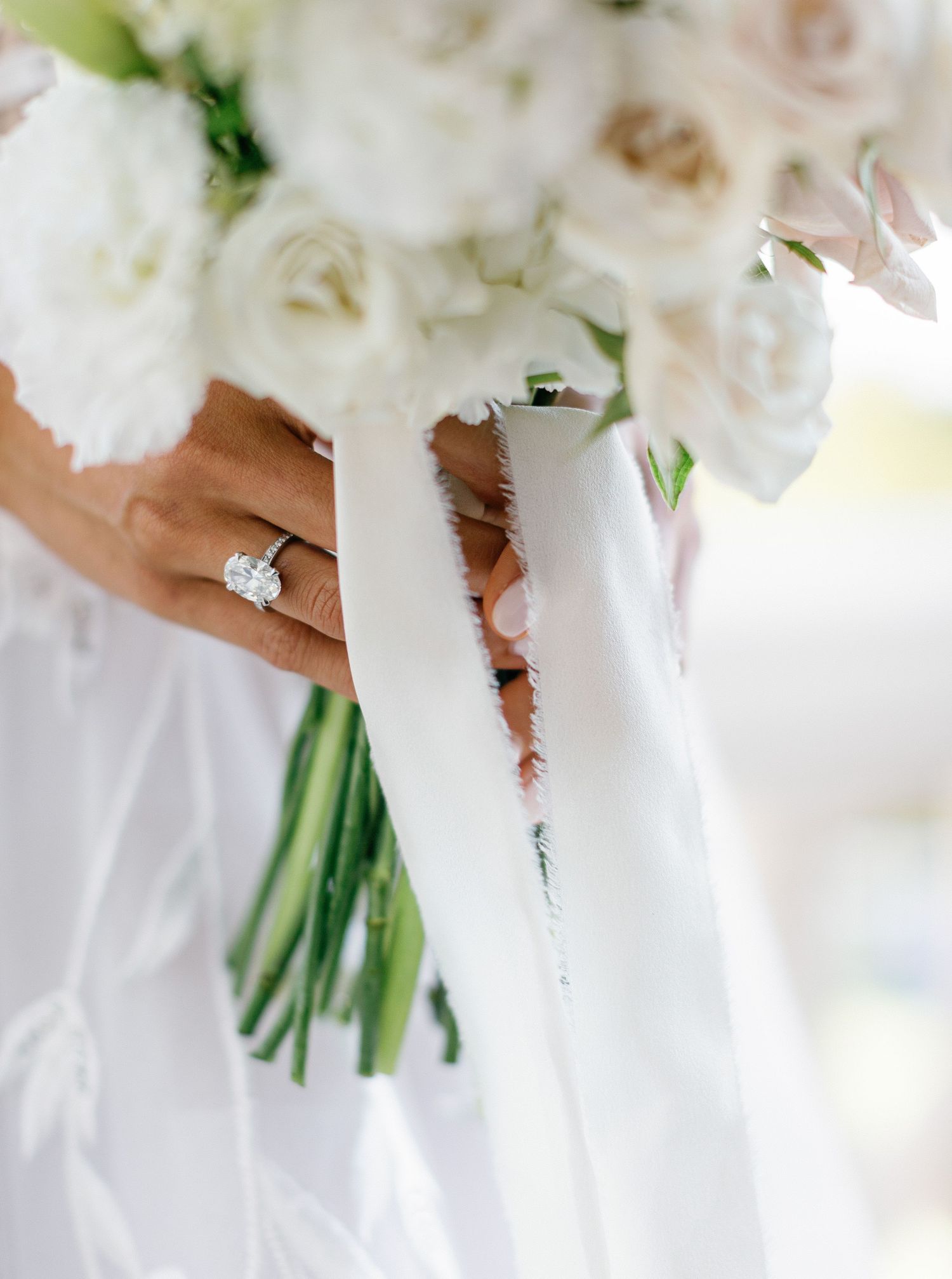 A close-up view of a white bridal bouquet and diamond engagement ring against a flowing white dress.