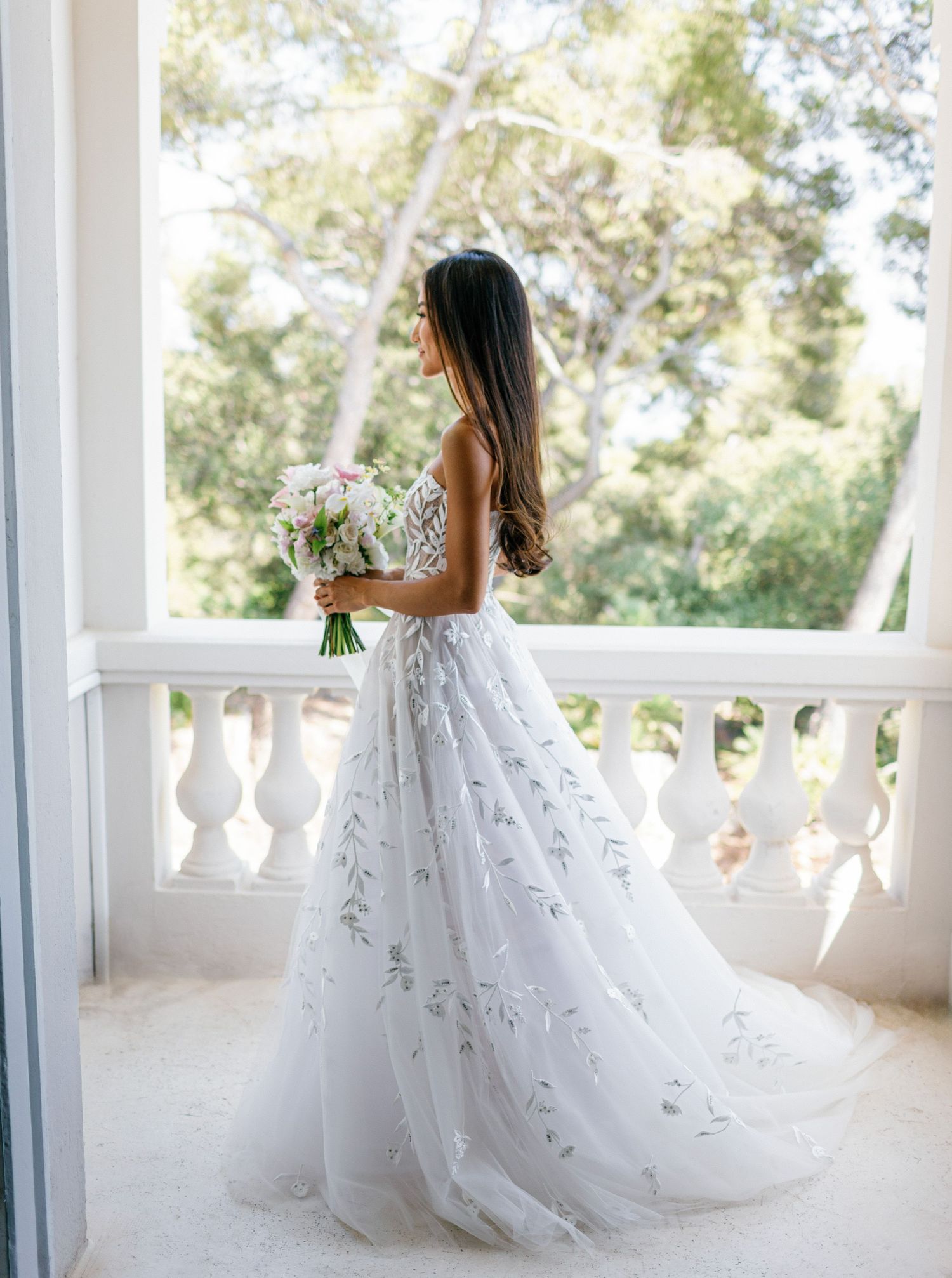 A bride in an elegant white wedding gown with floral details poses gracefully on a columned balcony with sunlit trees visible.