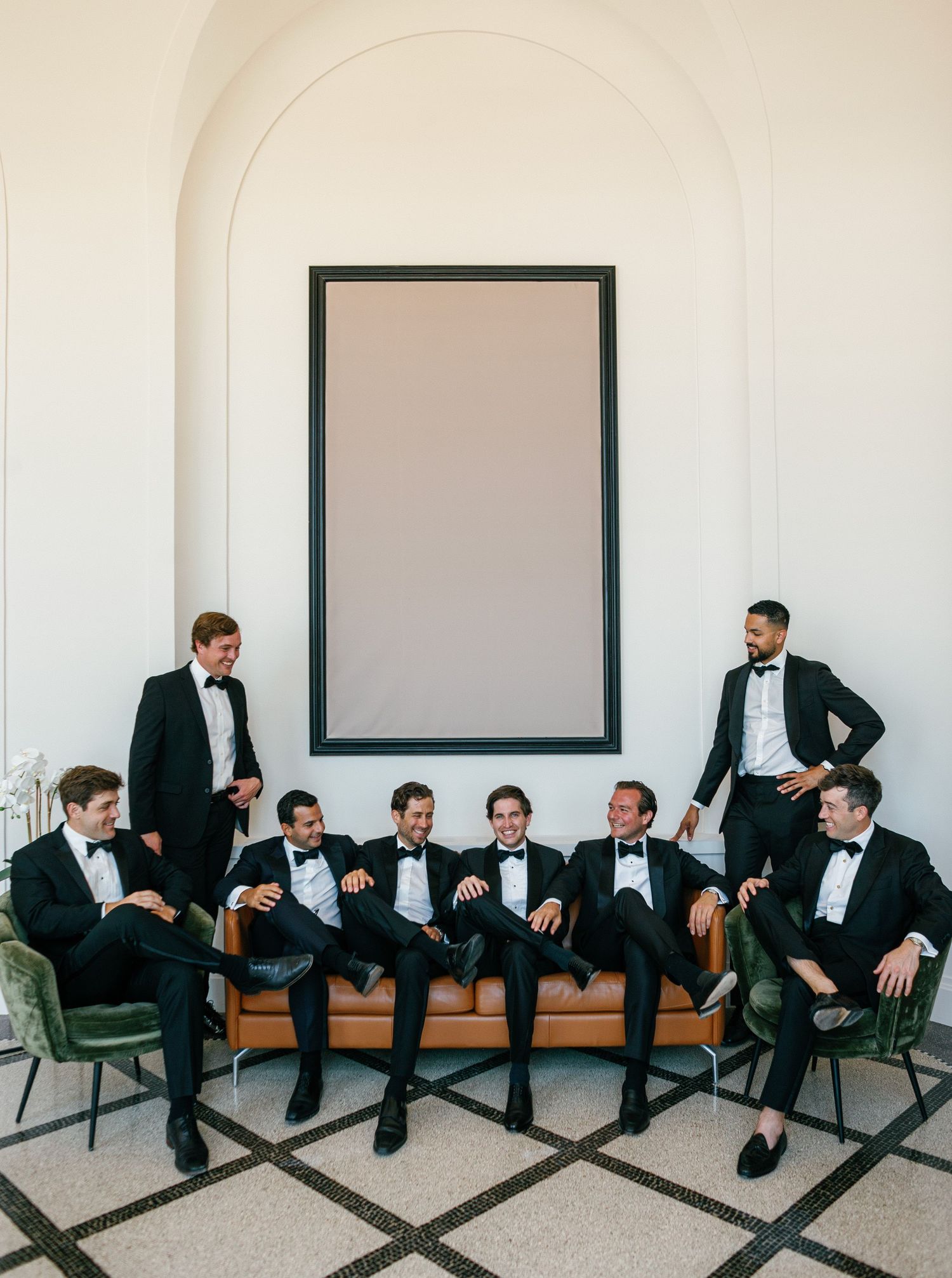 A creative panoramic sequence of people in formal attire lounging on chairs along a white wall with black frames.