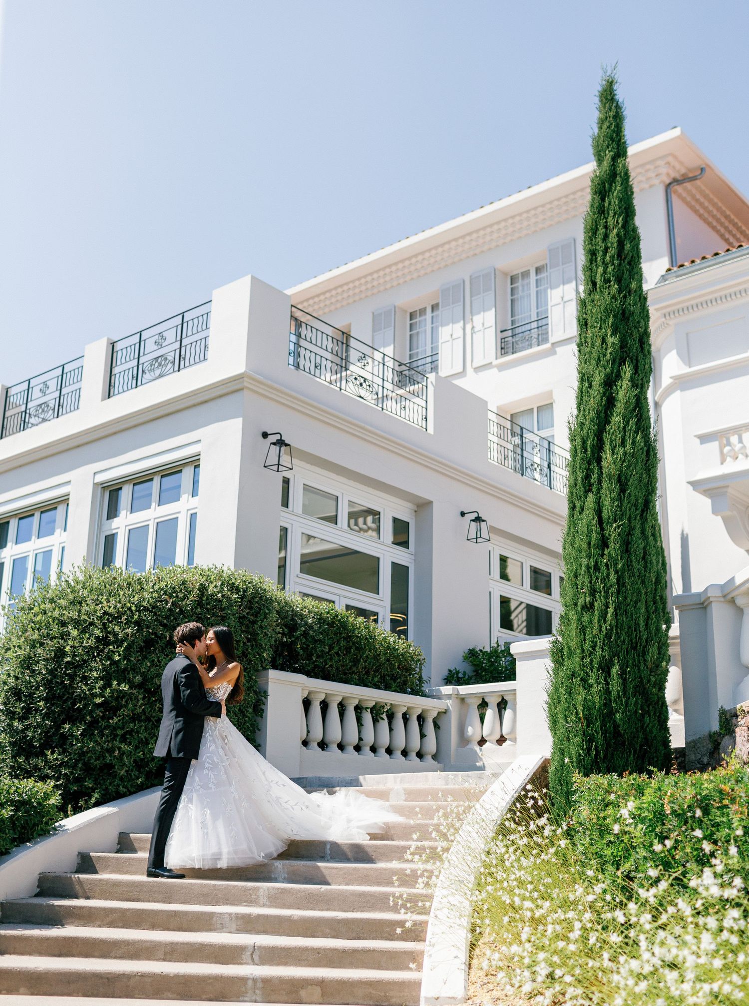 Newlyweds kissing on stairs surrounded by blooming flowers and cypress trees at Castel Bay, Hyères, French Riviera.
