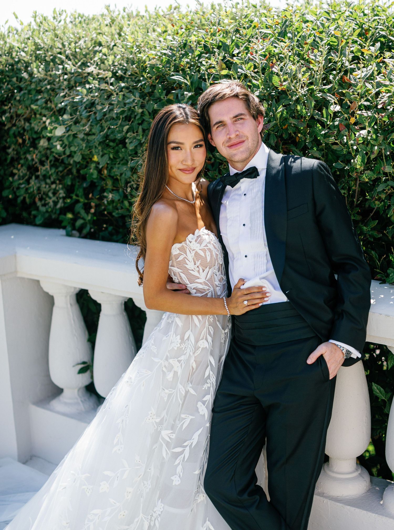 Wedding couple poses elegantly by white pillars and green hedges in outdoor garden setting.