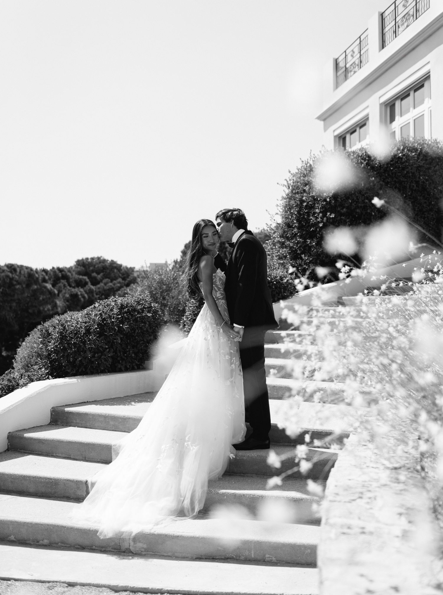Bride and groom kissing on stairs surrounded by blooming flowers at Castel Bay, Hyères, French Riviera.