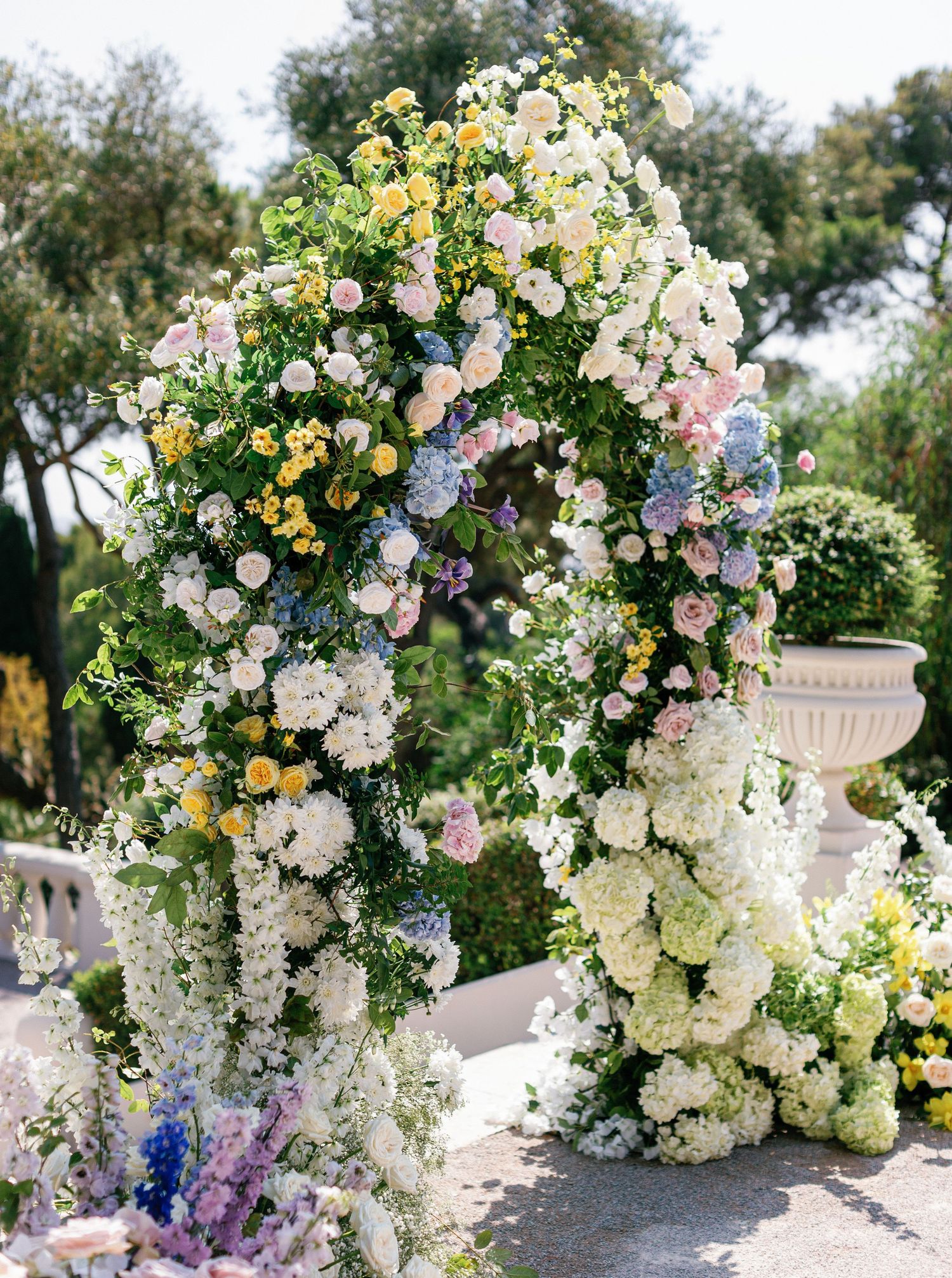 Elegant wedding floral arrangements featuring white, yellow, and purple blooms cascade beautifully in an outdoor garden setting.