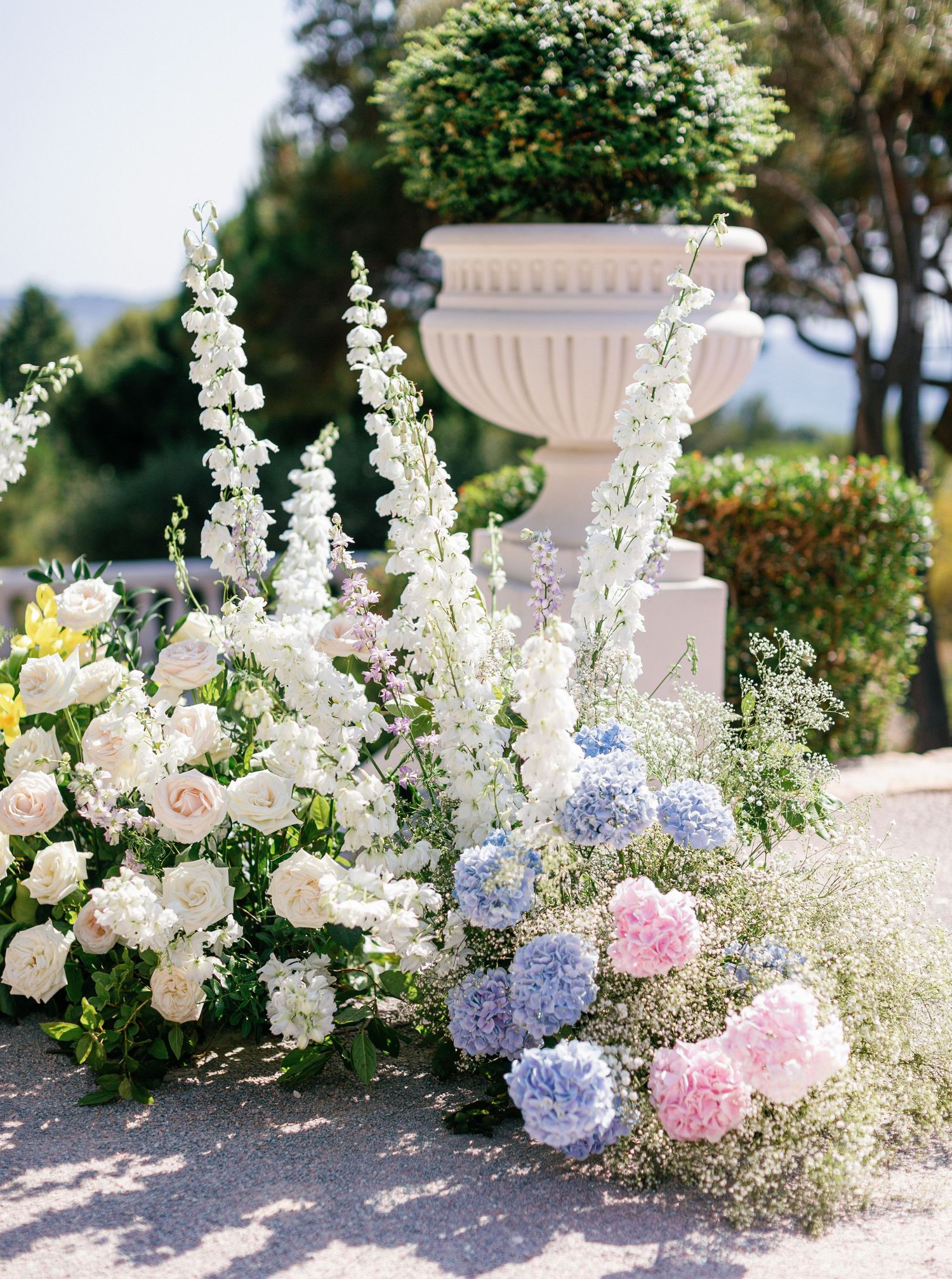 Elegant floral arrangements with white delphinium, pink and blue hydrangeas, and roses decorate an outdoor venue path.