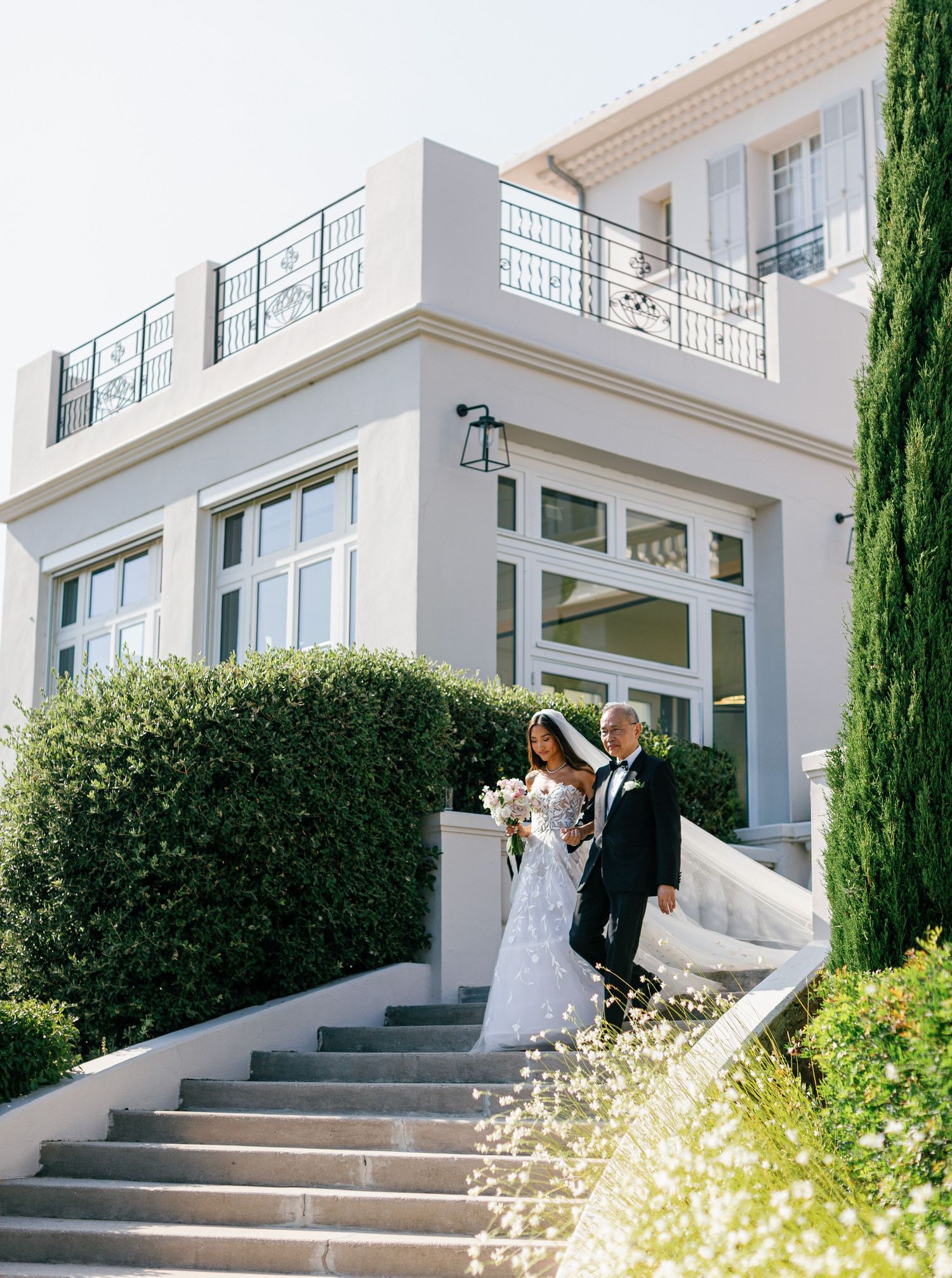 A romantic outdoor wedding photoshoot set on white stone stairs beside elegant cypress trees and a pristine white villa.