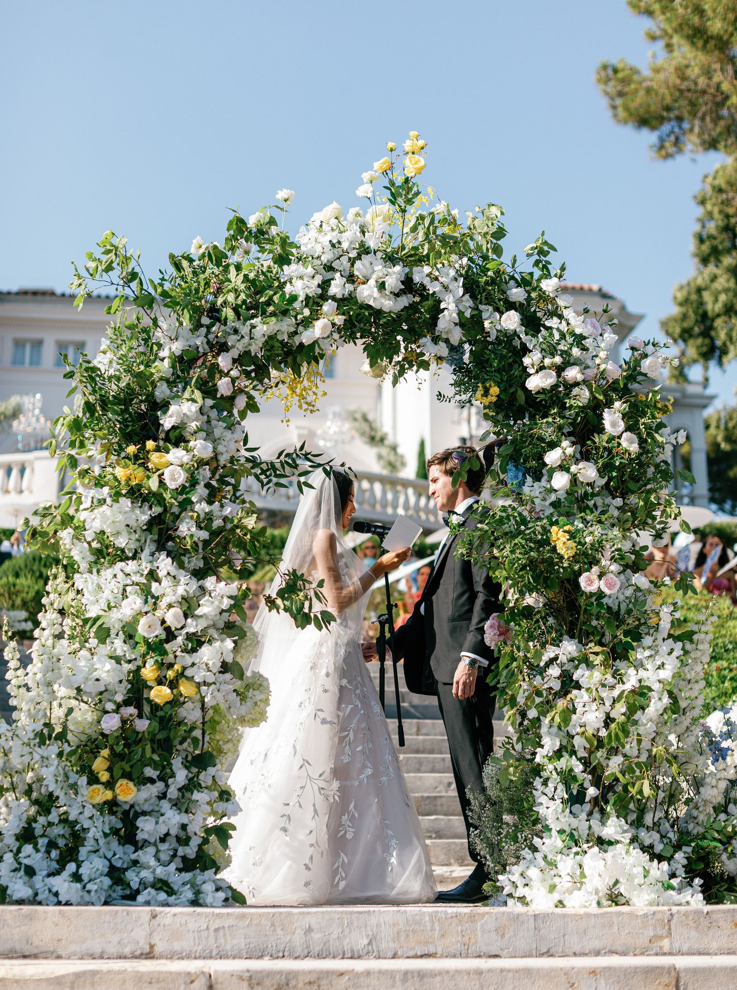 A wedding couple poses in front of an elegant white floral arch installation during the ceremony in a sunlit outdoor garden setting.