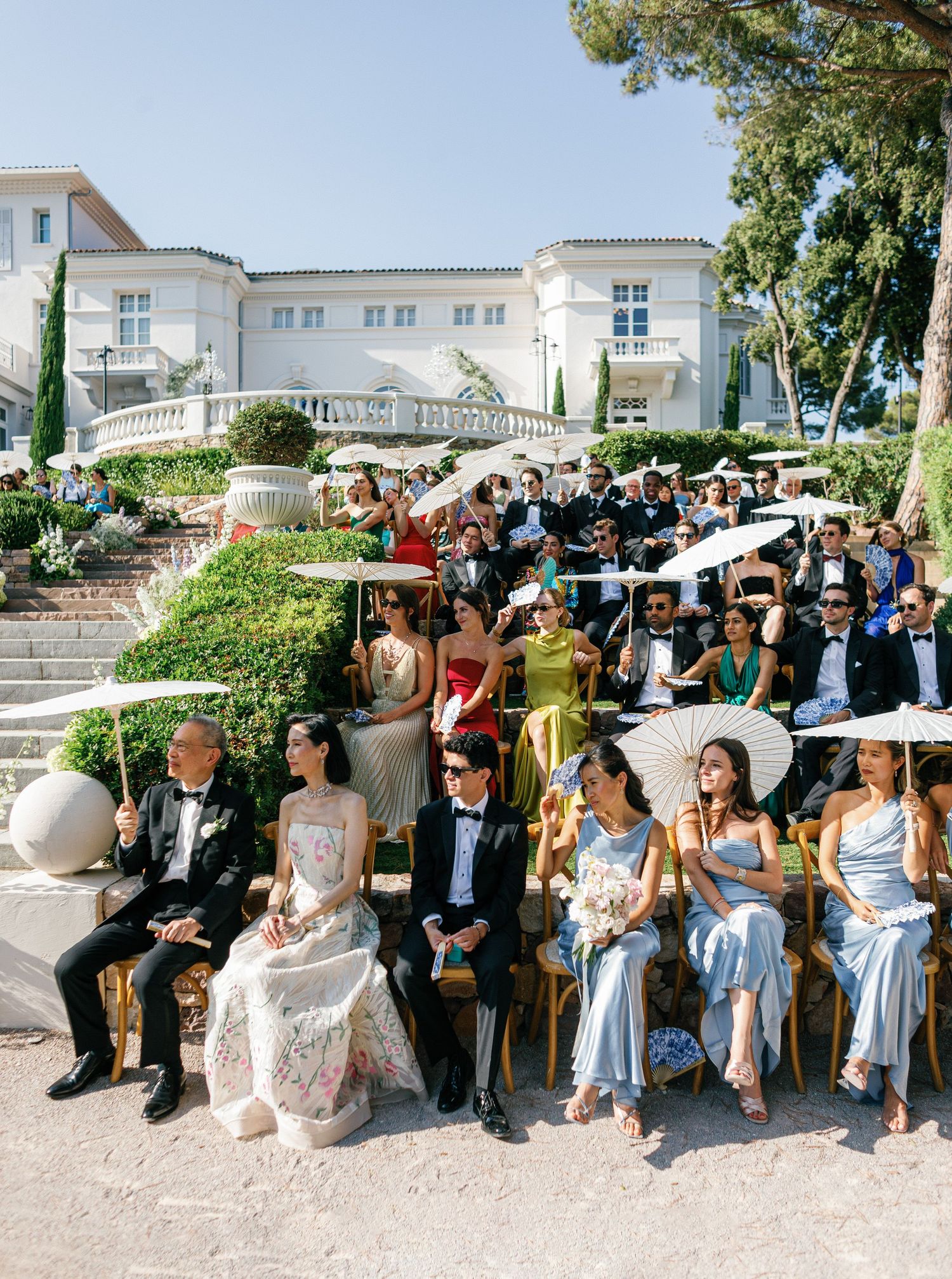 Wedding guests in formal attire sit outdoors on a sunny day at an elegant venue with white architecture.
