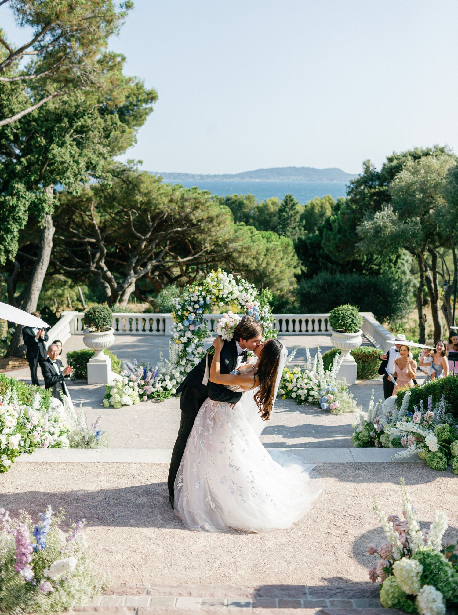 A romantic outdoor wedding ceremony with elegant white floral arch, garden decor and sea views in the background.