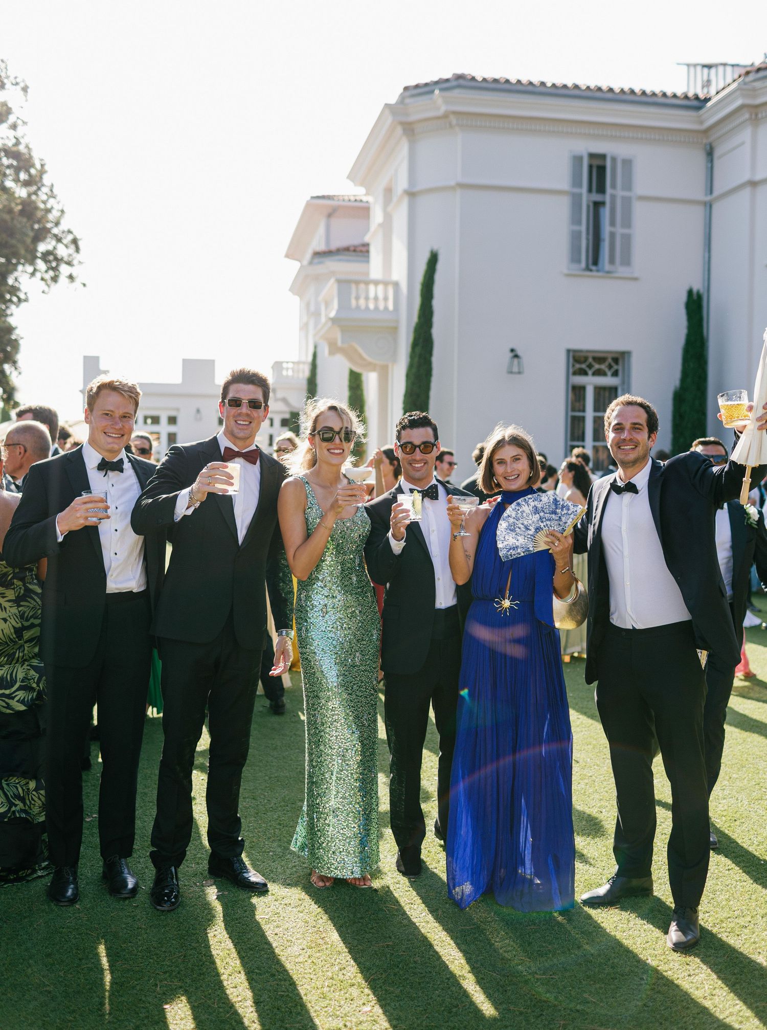 Wedding party in formal attire raises champagne glasses for a celebratory toast on a sunny lawn outside at Castel Bay, Hyères.