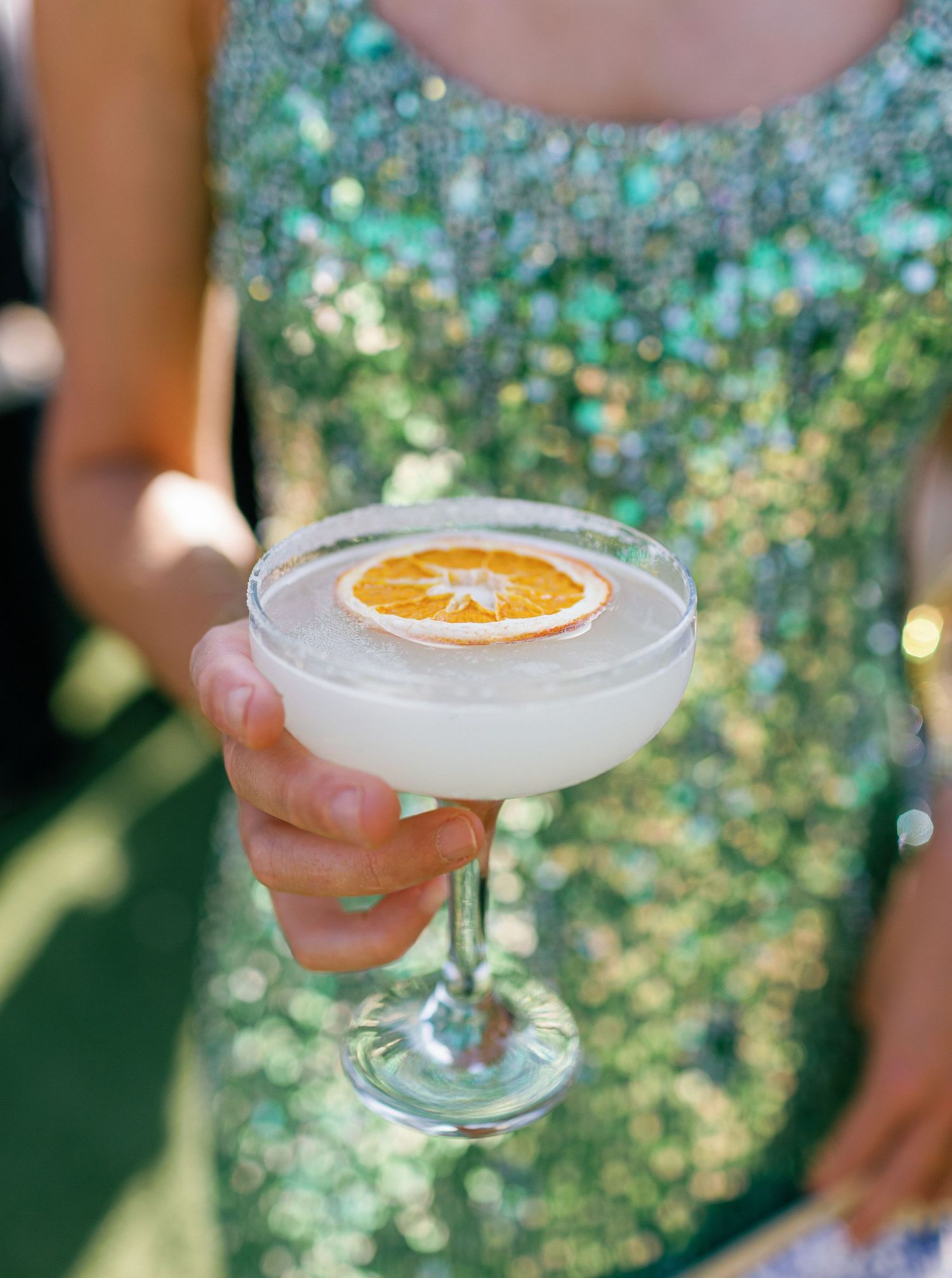 A hand holds an elegant coupe glass with a citrus cocktail garnished with orange slice against a sunlit bokeh background.
