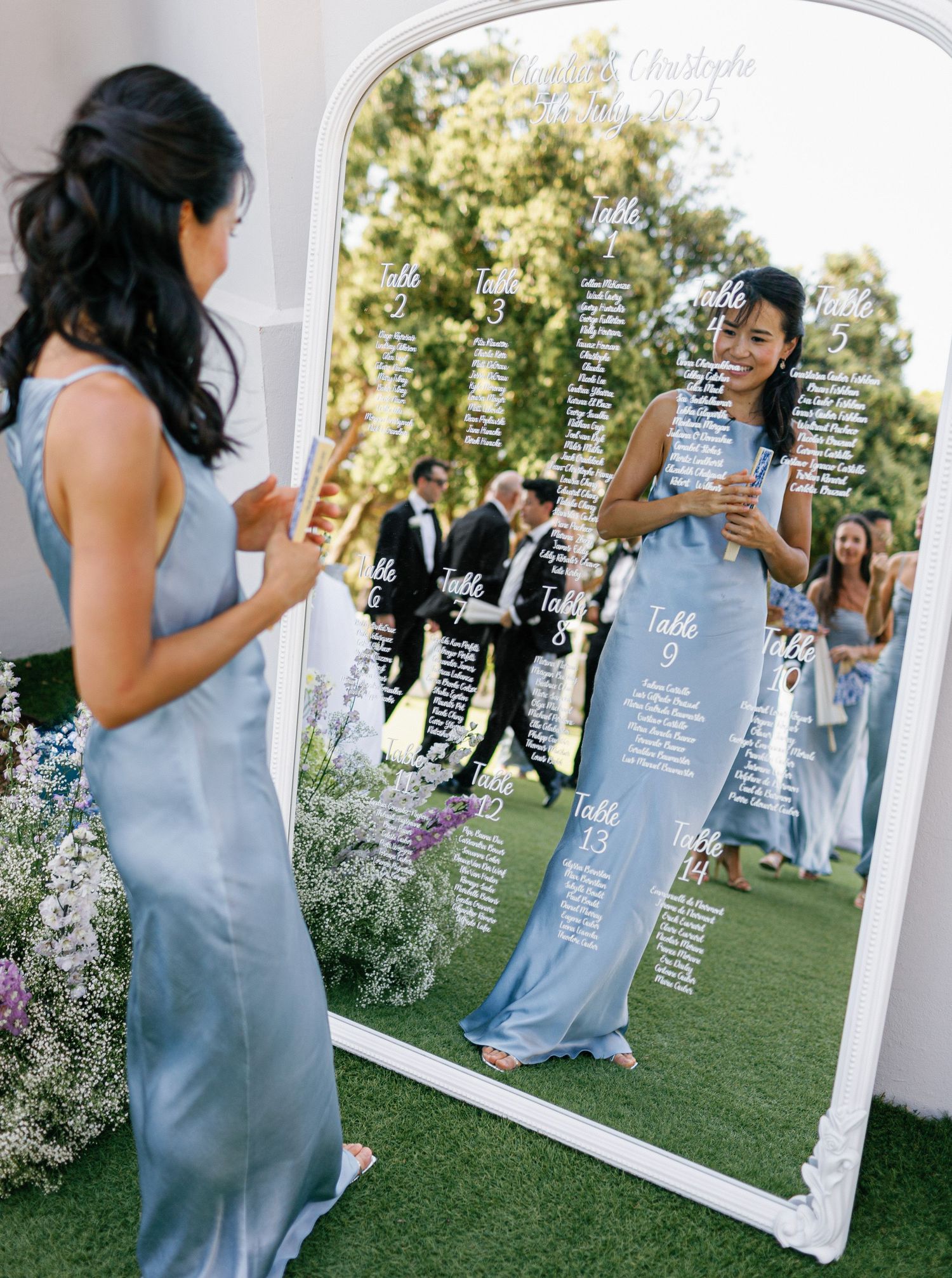A mirrored seating chart display at an outdoor wedding shows guest names reflected against a floral garden setting.