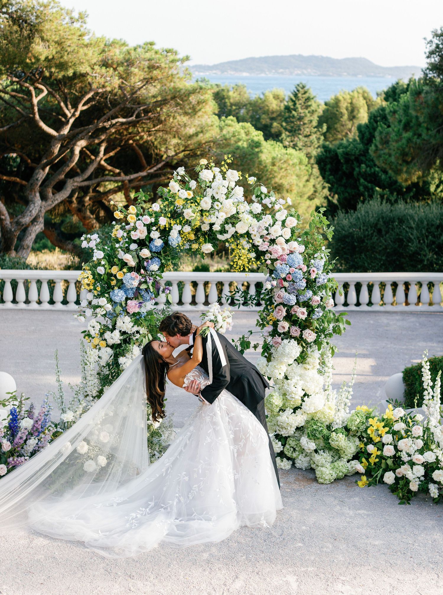 Bride and groom kissing under floral arch against scenic sea backdrop at Castel Bay, Hyères, French Riviera.