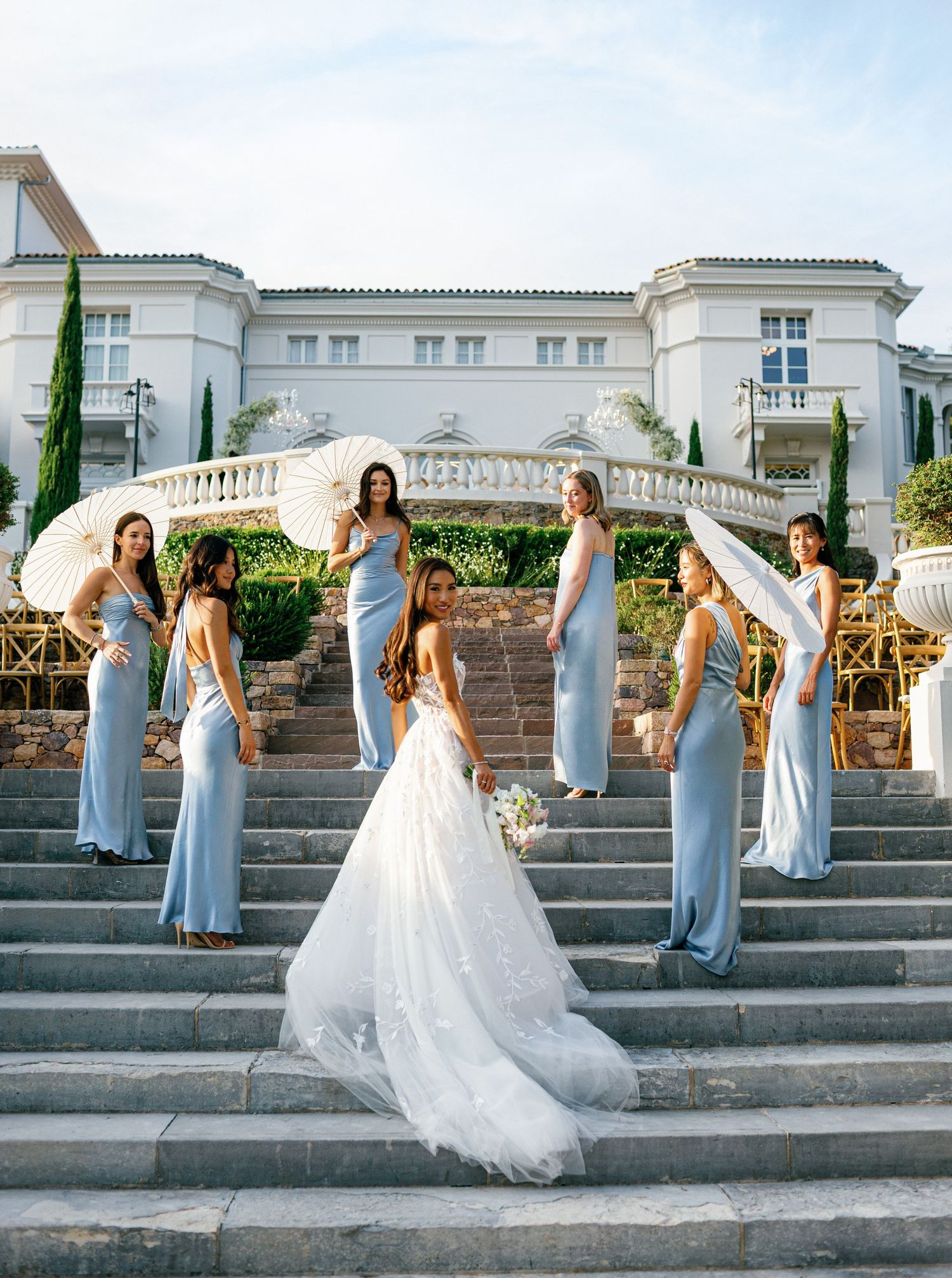 Bride and bridesmaids in light blue dresses pose together on elegant white stone steps of a mansion during golden hour.
