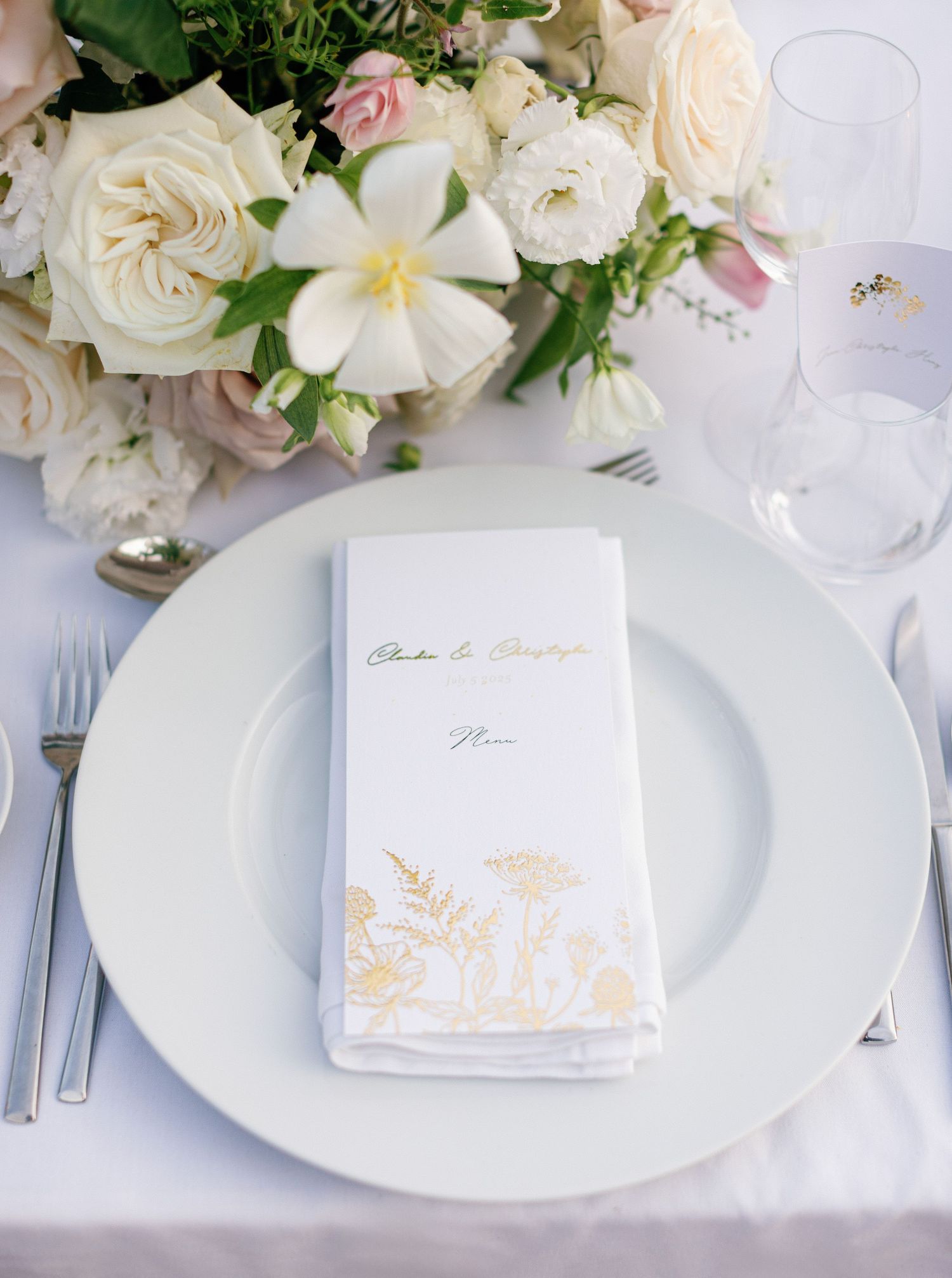 Elegant wedding place setting with white linen napkin, white roses centerpiece, and gold foil menu card on pale blue plate.