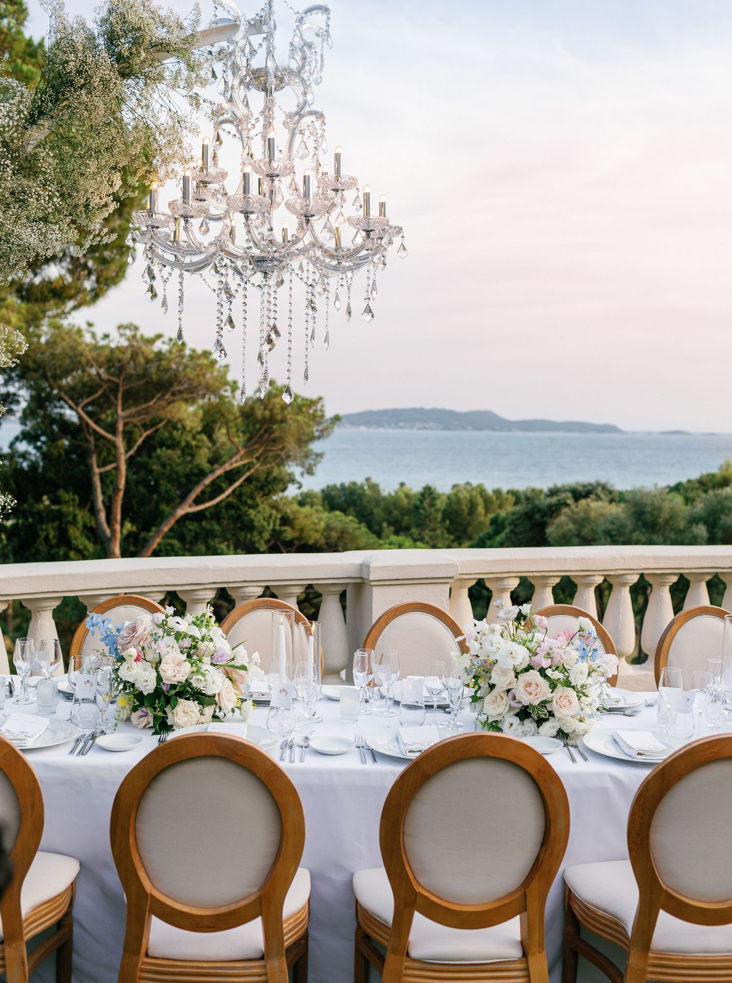 Elegant wedding reception table setup with crystal chandeliers and panoramic ocean views through arched windows at sunset.