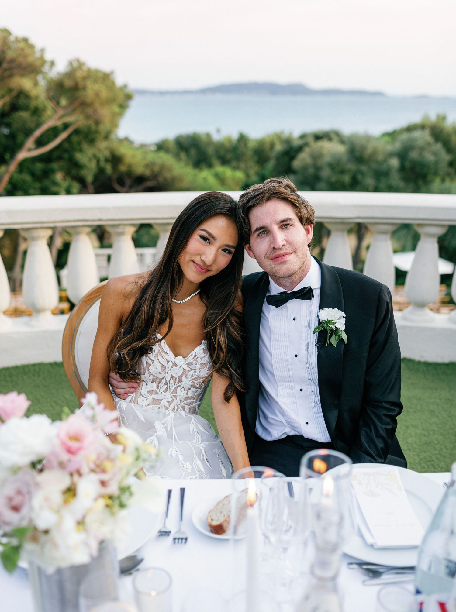 Elegant wedding reception table setup outdoors with pink florals and white tablecloths against a scenic sea backdrop.