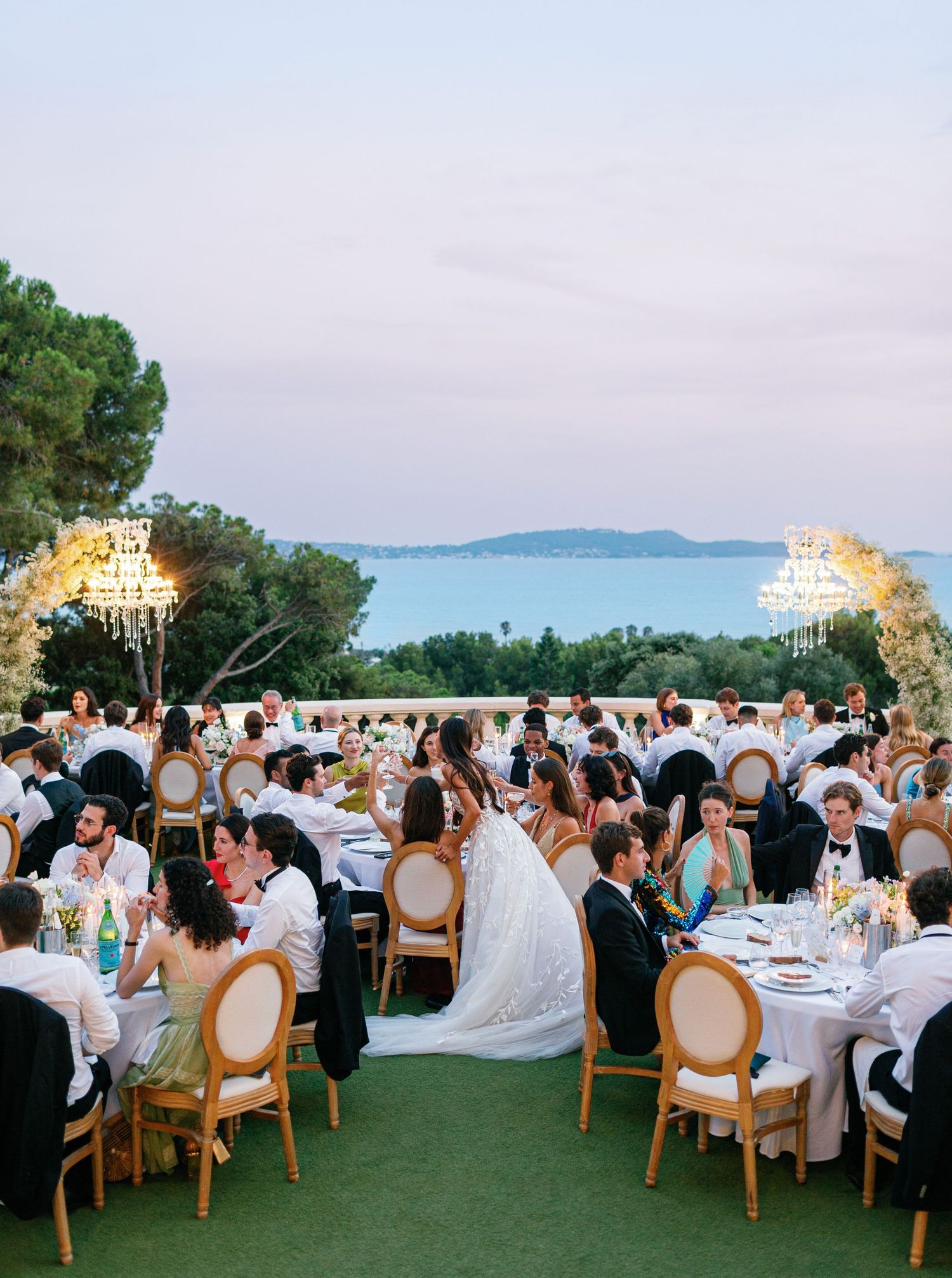 Elegant outdoor wedding reception with round tables on green lawn overlooking ocean view at dusk with hanging chandeliers at Castel Bay.
