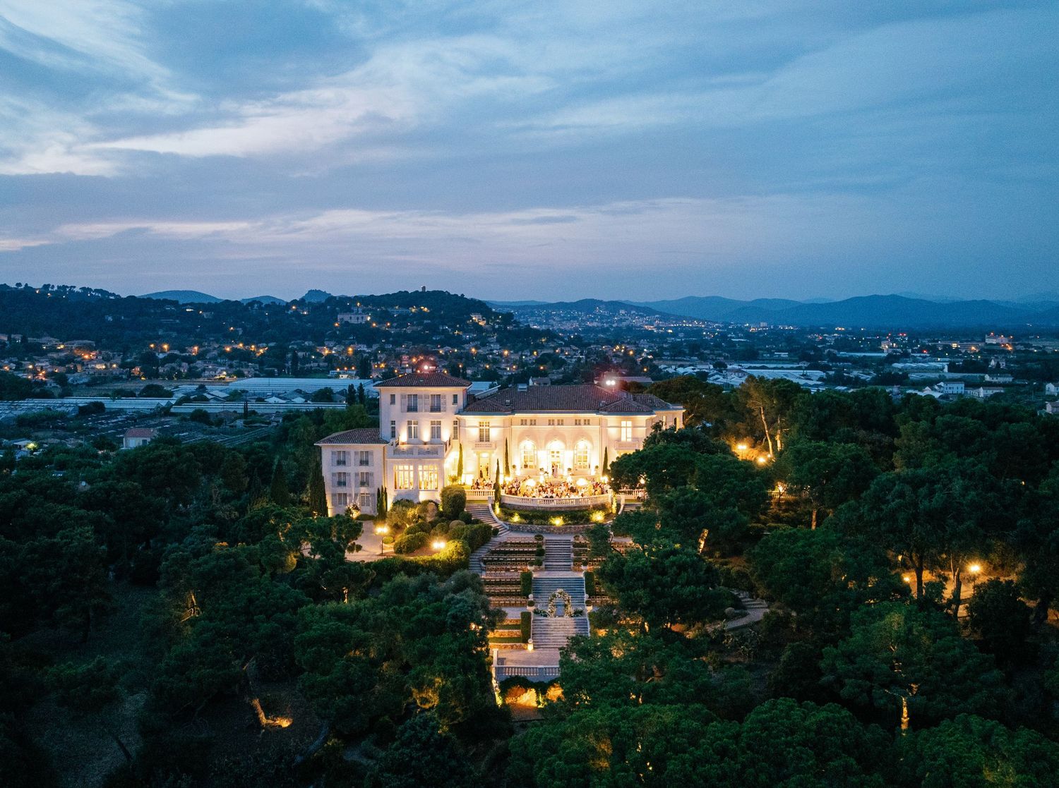 Panoramic sunset view of an illuminated luxury villa Castel Bay nestled in lush hillside overlooking a Mediterranean coastal town.
