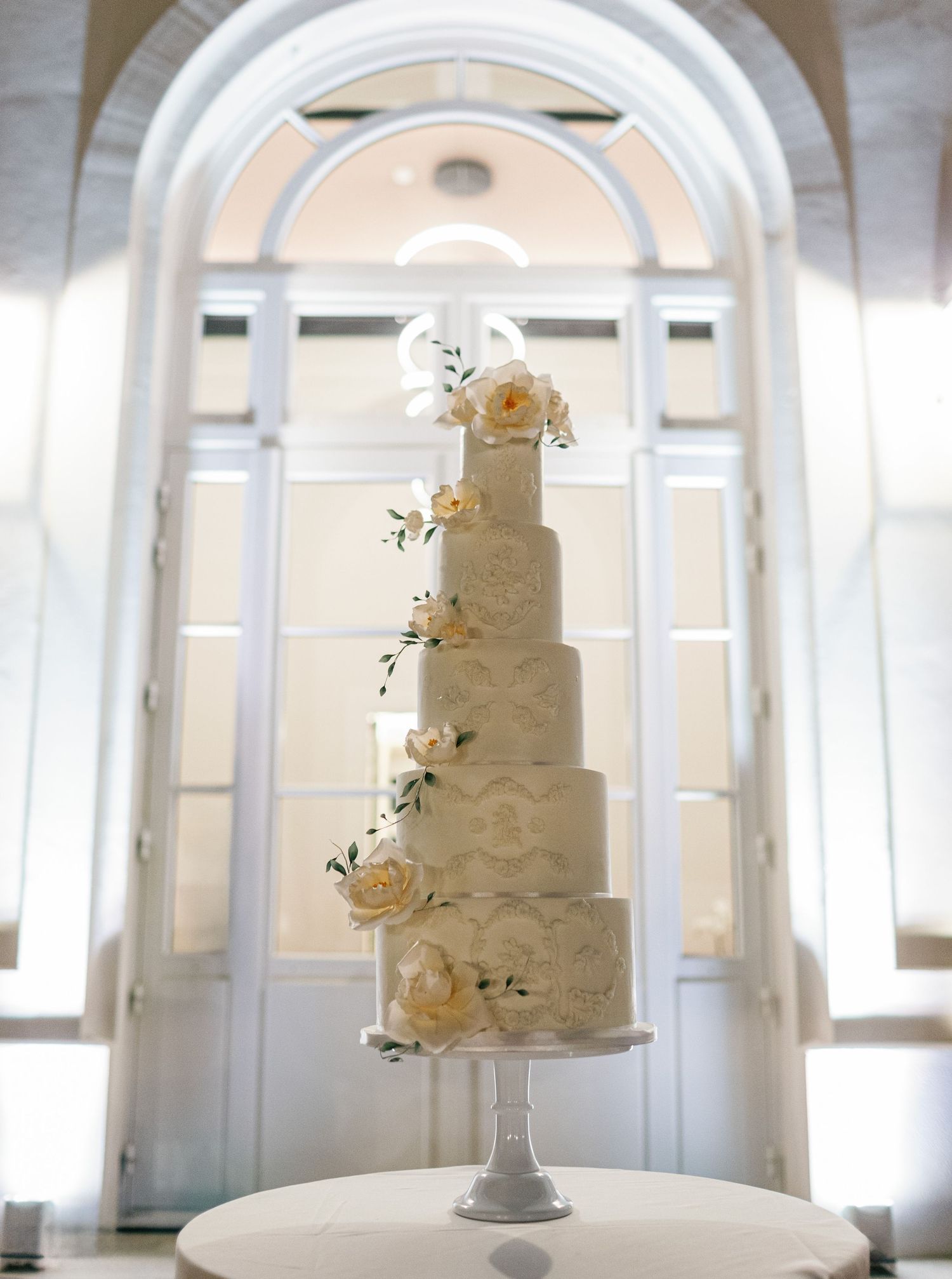 Elegant three-tiered white wedding cake with delicate peach roses displayed in classic arched venue setting.