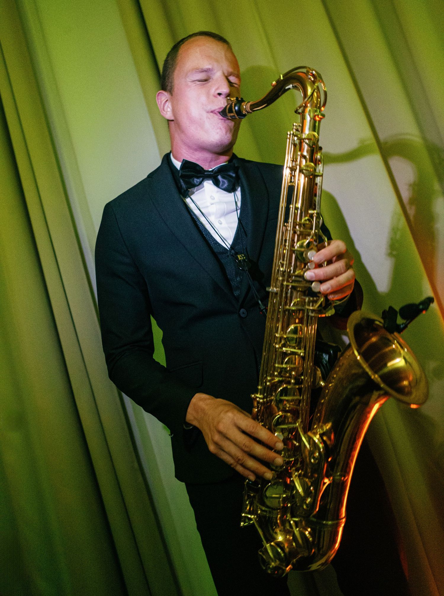 Professional musician performs with a brass saxophone against a green illuminated stage backdrop creating moody ambiance.