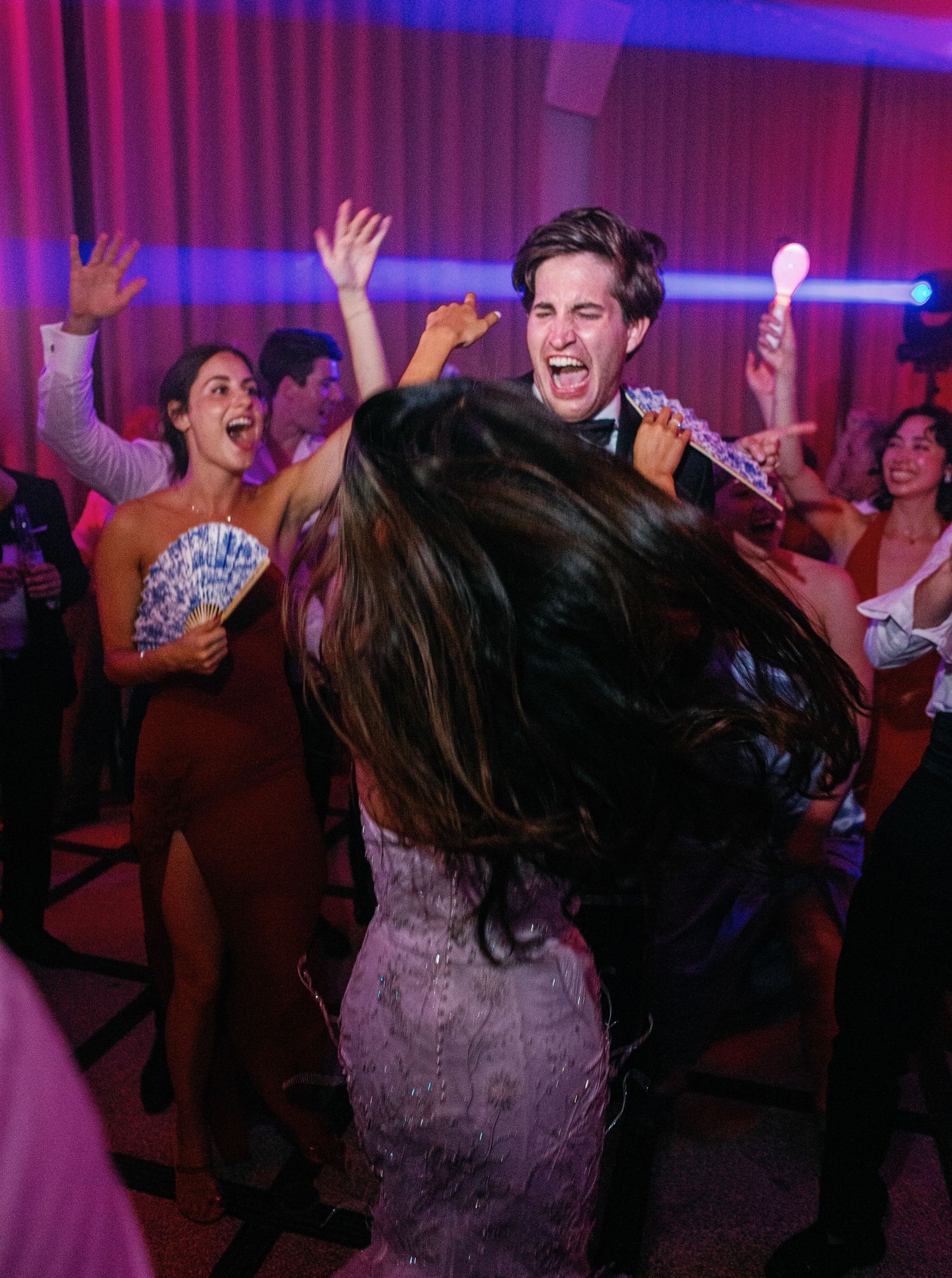 Wedding guests dancing and celebrating with energetic movements in purple and blue lighting at reception party.