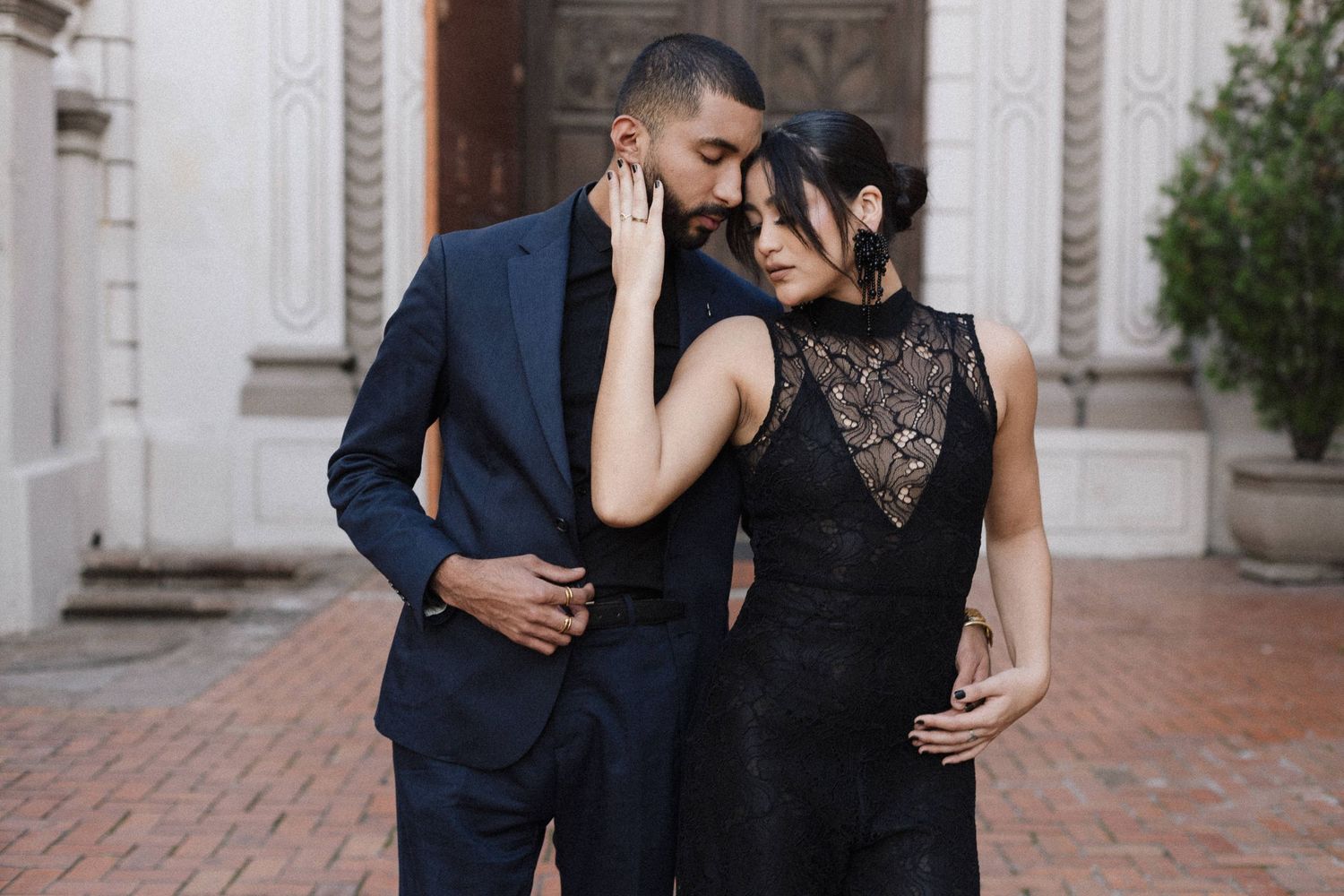 Elegant couple in formal attire embrace against ornate architectural backdrop.