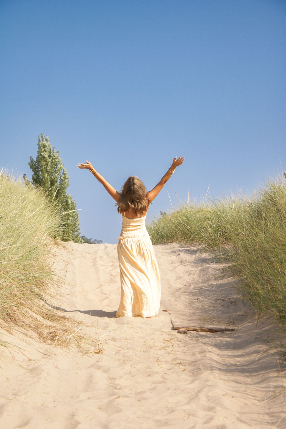 Figure in light colored dress stands joyfully atop sandy beach dunes with arms raised.