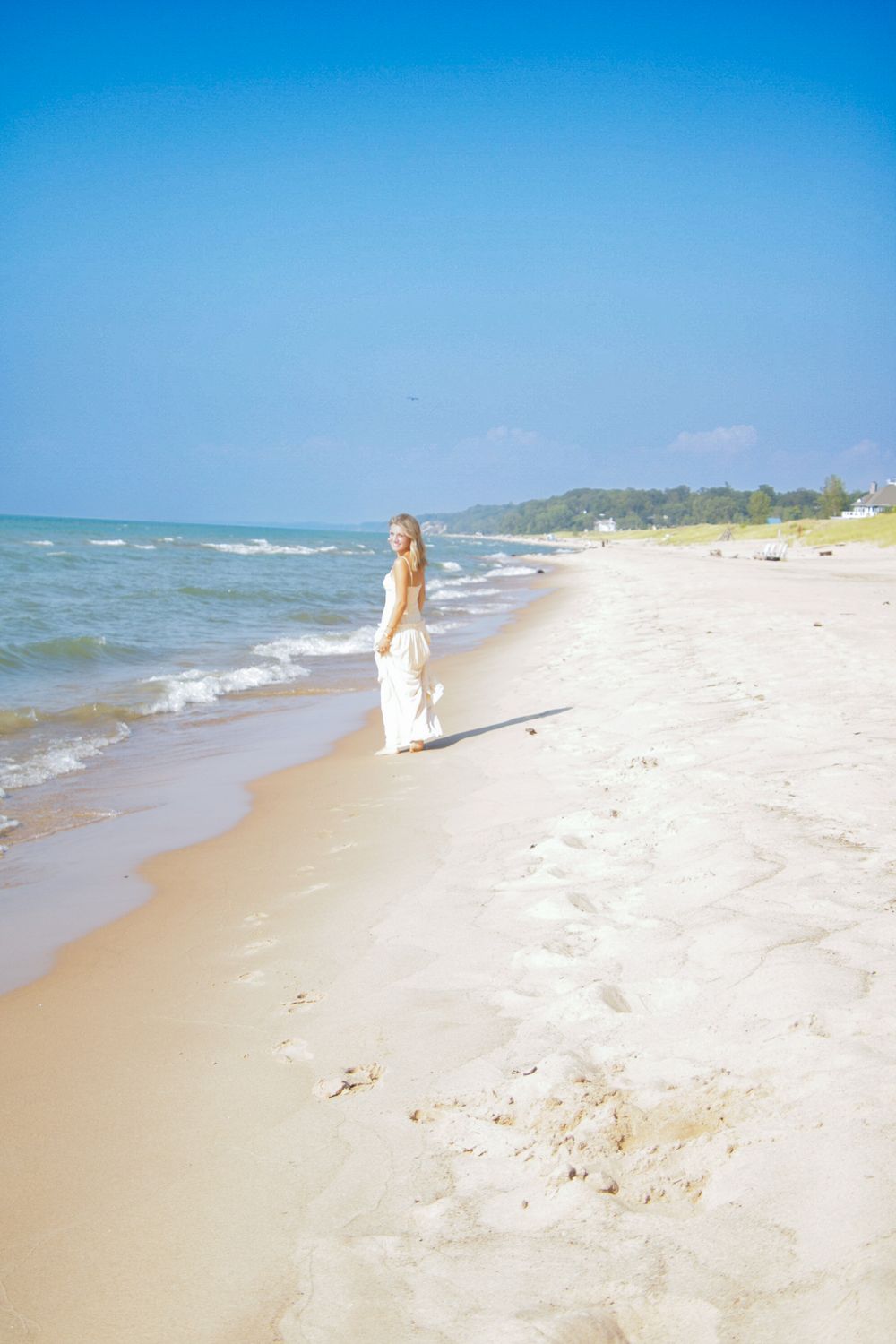 Figure in a white dress walking along a peaceful beach shoreline with blue water and clear skies.