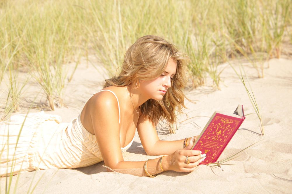 Person relaxing on beach sand while reading a red book among coastal dune grass.