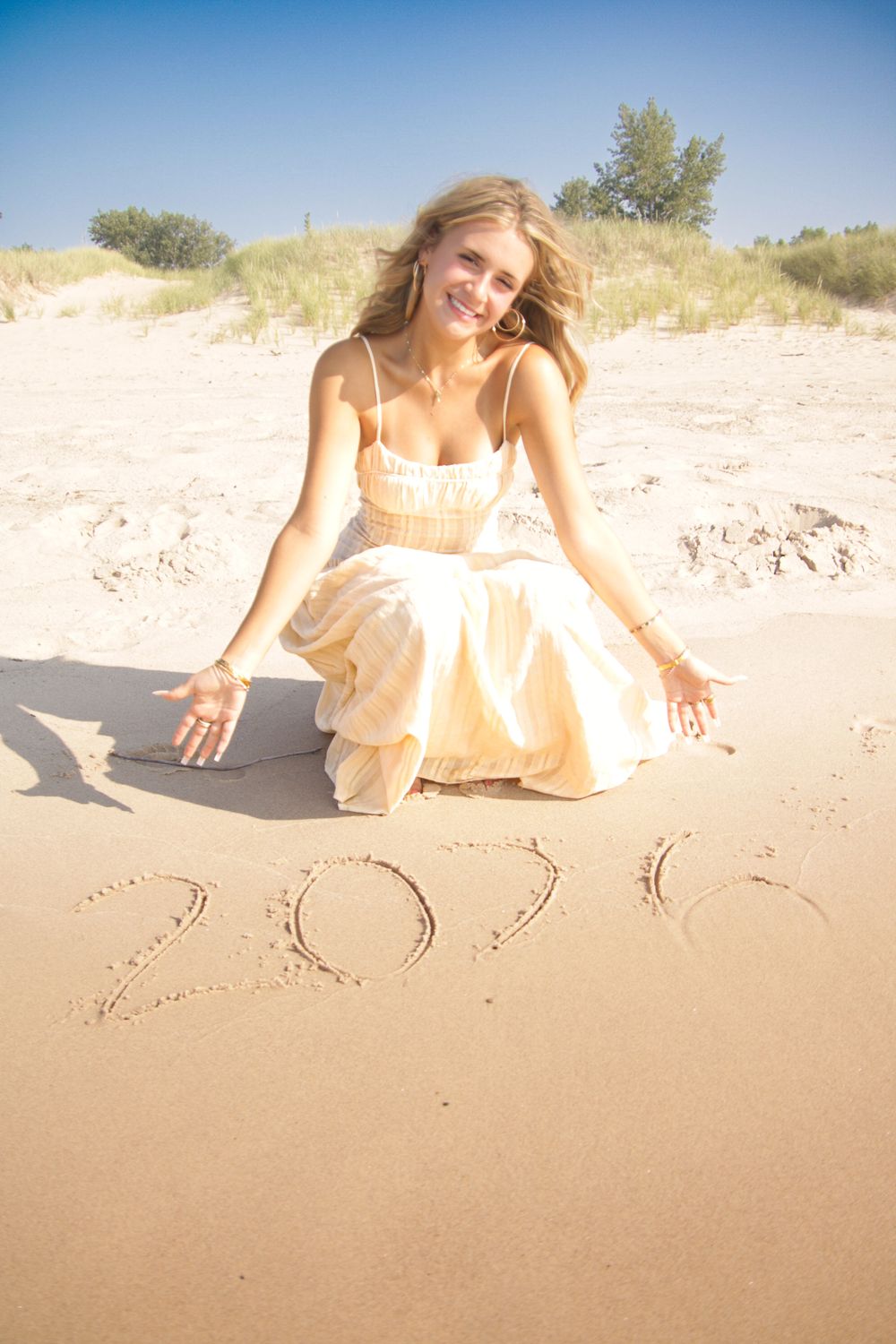 Person in light dress sitting by '2011' written in beach sand with dunes in background.